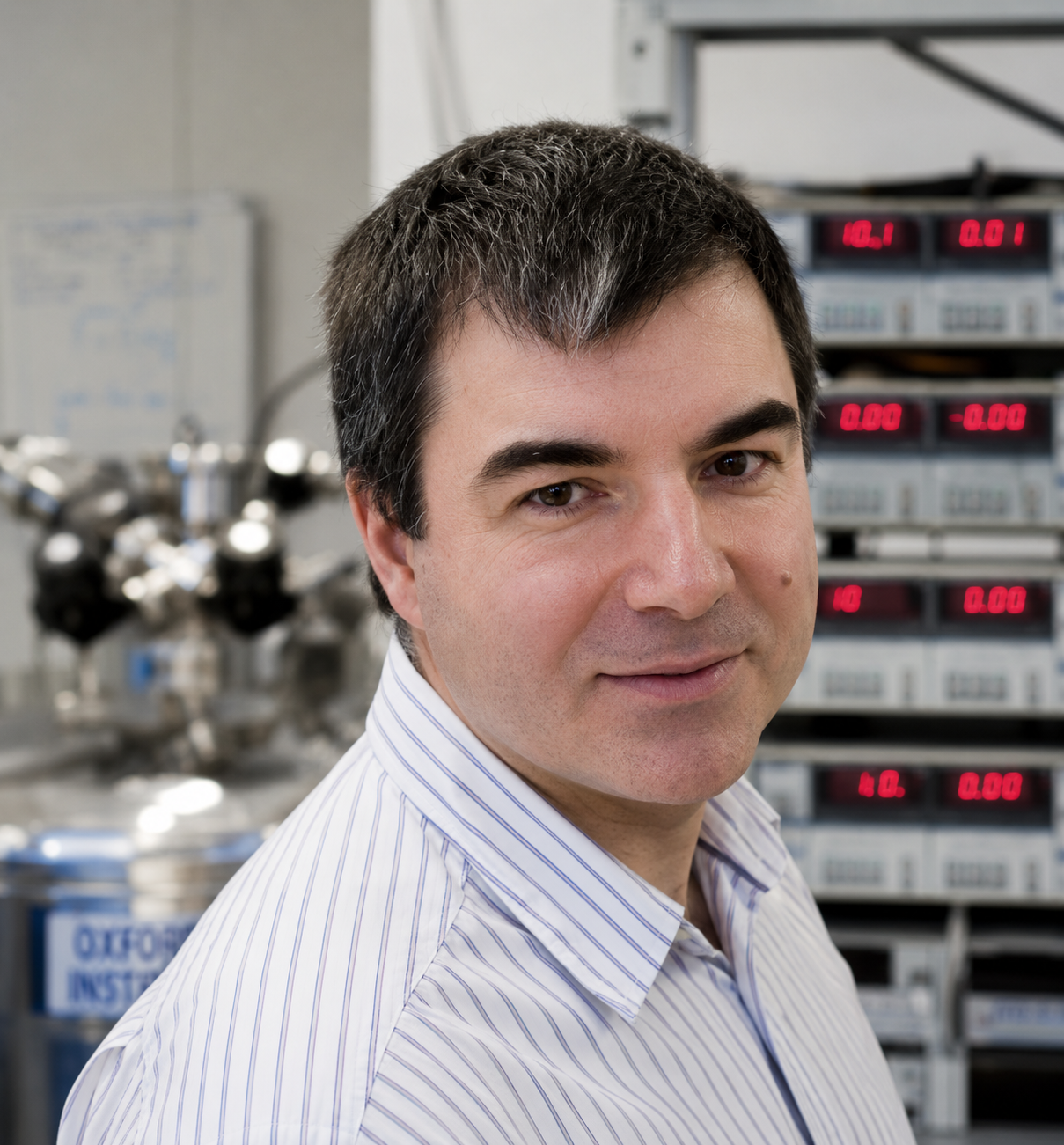 A man in a light blue shirt smiling, with laboratory equipment visible in the background.